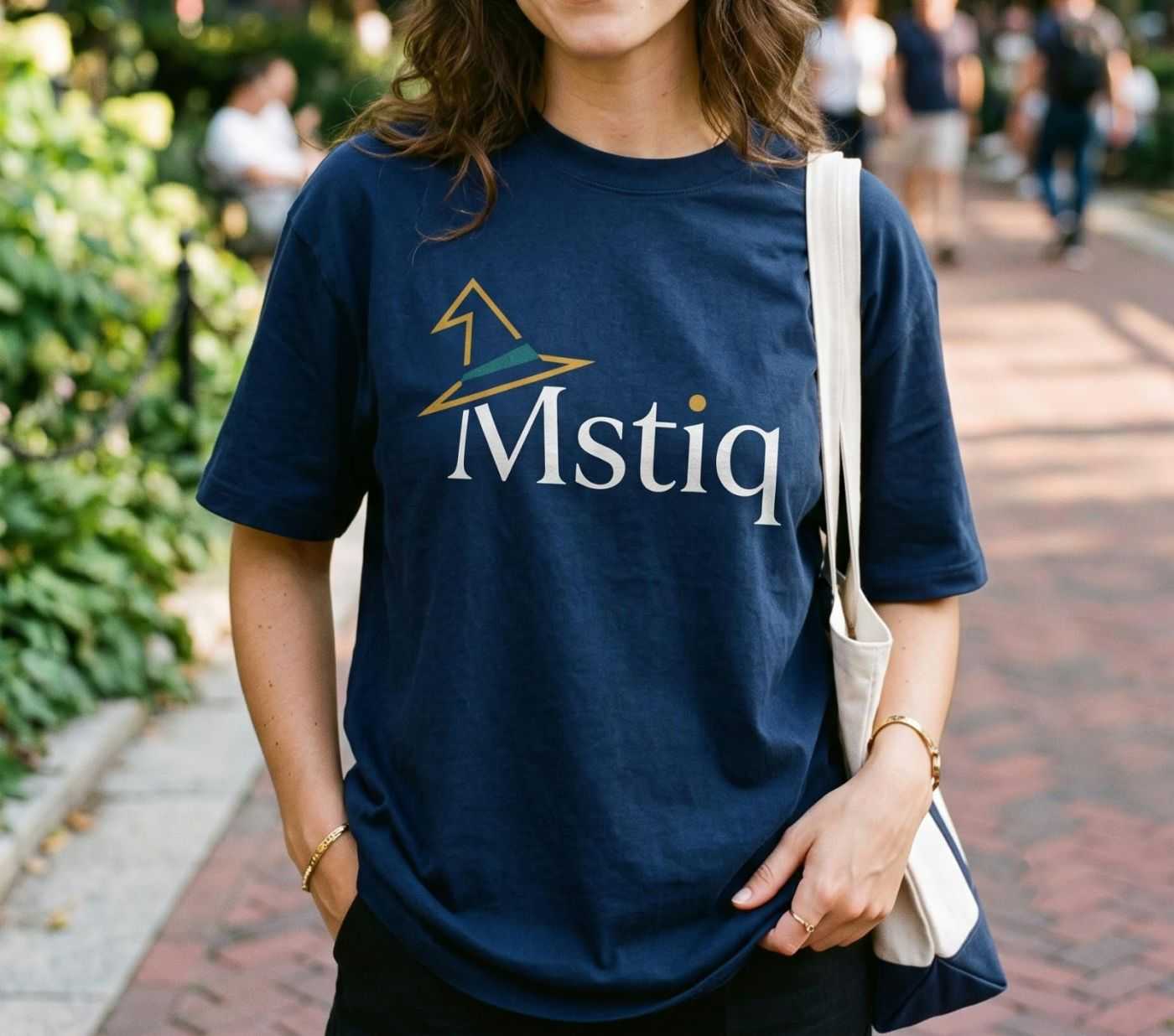 A person walking through a park wearing a navy Mstiq t-shirt and carrying a canvas tote bag